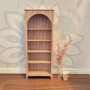 May include: A miniature wooden bookshelf with an arched top and four shelves. The bookshelf is made of light-colored wood and is empty. A small vase with dried flowers and a small potted plant are next to the bookshelf.