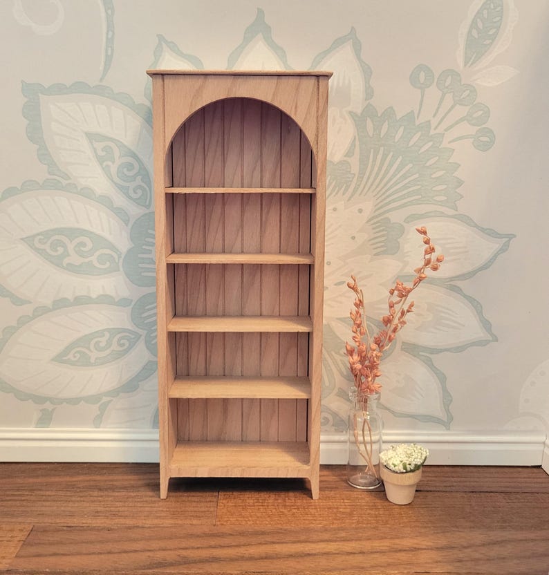 May include: A miniature wooden bookshelf with an arched top and four shelves. The bookshelf is made of light-colored wood and is empty. A small vase with dried flowers and a small potted plant are next to the bookshelf.