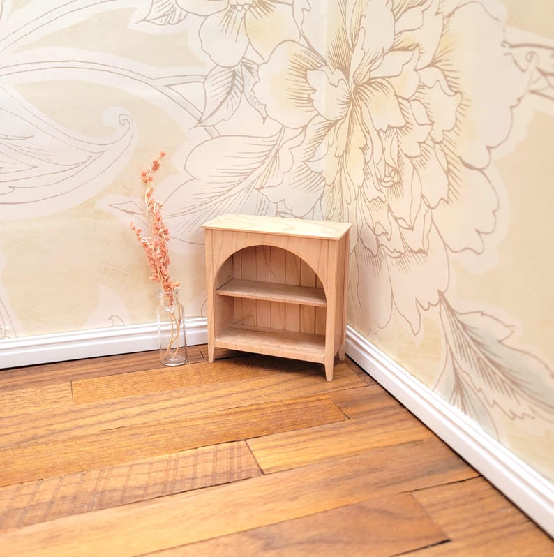 May include: A miniature wooden bookshelf with two shelves and an arched top. The small cabinet is made of light-colored wood and is designed for a dollhouse or miniature setting. A small glass vase with pink flowers is next to the bookshelf.