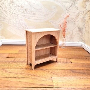 May include: A miniature wooden bookshelf with two shelves and an arched opening. The bookshelf is made of light-colored wood and has a simple, classic design. It is a dollhouse-sized piece of furniture.