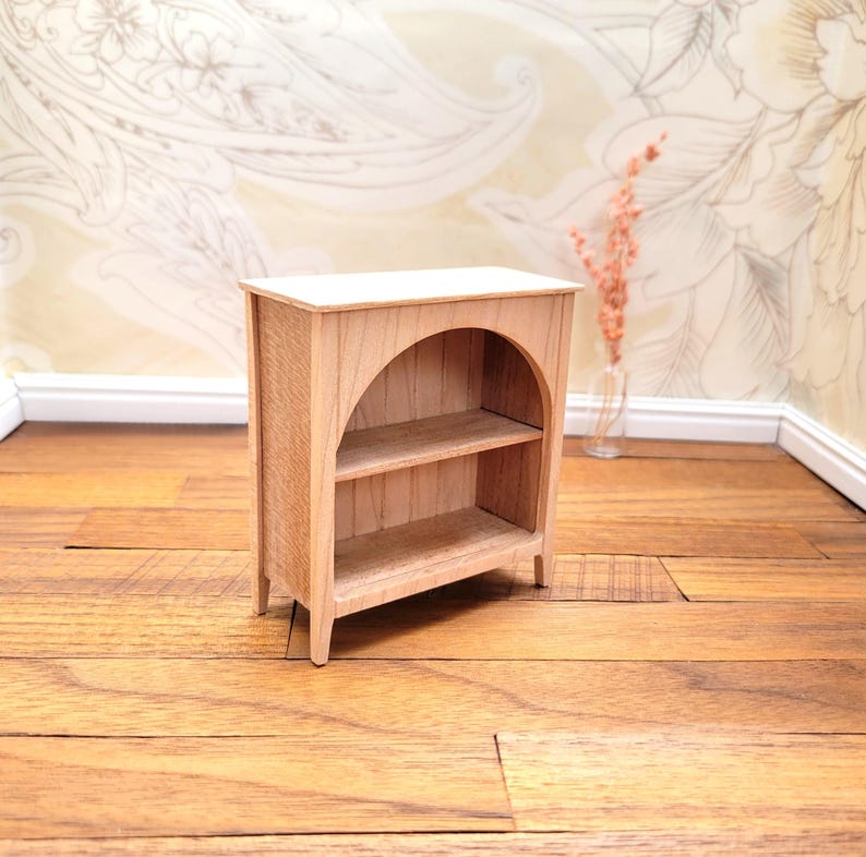 May include: A miniature wooden bookshelf with two shelves and an arched opening. The bookshelf is made of light-colored wood and has a simple, classic design. It is a dollhouse-sized piece of furniture.