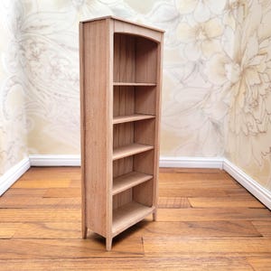 May include: A miniature wooden bookshelf with four shelves. The bookshelf is made of light-colored wood and has a curved top. It stands on a wooden floor, and the background features a floral wallpaper.