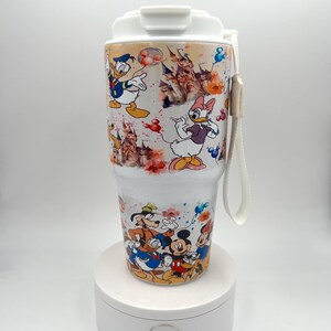Könnte beinhalten: Ein weißer Reisebecher mit Disney-Figuren wie Donald Duck, Daisy Duck, Mickey Mouse und Goofy. Der Becher hat einen weißen Deckel und einen passenden Riemen. Das Design umfasst Bilder eines Schlosses und farbenfrohe florale Akzente.