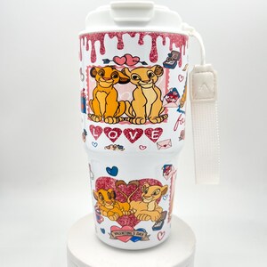Könnte beinhalten: Ein weißer Reisebecher mit einem Valentinstags-Design, das Löwenjunge, Herzen und das Wort "LOVE" zeigt. Der Becher hat einen weißen Deckel und einen weißen Riemen. Das Design enthält den Text "VALENTINE'S DAY".