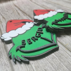 May include: Green Christmas ornament with a red Santa hat and the name "Sergio" in black letters. The ornament has a black border and a white trim on the hat. The ornament is made of wood and is on a wooden surface.