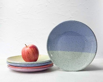 May include: A stack of four round ceramic plates with a speckled finish in shades of light blue, pink, and cream, topped with a single red apple. Beside the stack stands one additional round ceramic plate featuring a two-tone design of blue and light green with a speckled texture throughout the surface.