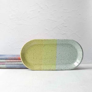 May include: An oval ceramic tray featuring a speckled finish with a color-blocked design. The left half is a bright lime yellow, while the right half is a soft mint green. A stack of similar trays in various pastel colors sits to the left.