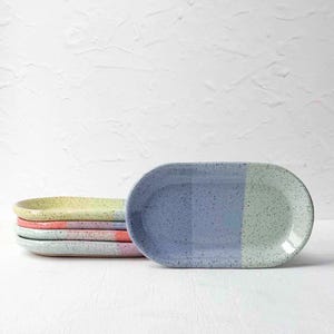 May include: A stack of four oval ceramic dishes with a speckled finish. Each dish features a color-blocked design with two distinct pastel hues, including shades of yellow, pink, blue, and green, arranged in a horizontal orientation.