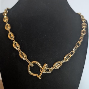 May include: A gold chain necklace with a large, oval-shaped link design. The necklace has a unique, hook-style clasp.