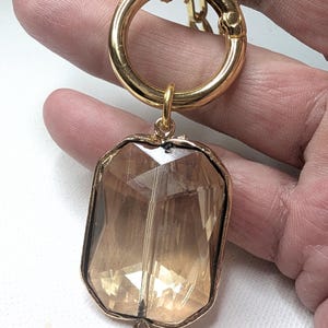 May include: A gold-colored keychain with a large, faceted, rectangular amber-colored pendant. The pendant is encased in a gold-colored metal frame. The keychain has a large, round clasp and a smaller ring connecting the pendant.