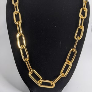 Op de afbeelding: Een gouden ketting met grote, rechthoekige schakels. De ketting wordt getoond op een zwarte fluwelen standaard.