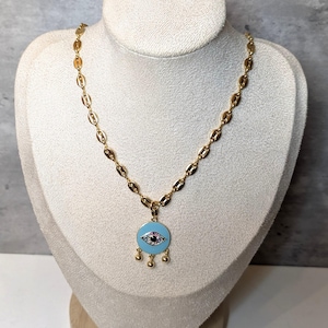 May include: A gold-colored necklace with a chain of oval links. The pendant is a light blue circle with an eye design and three small gold-colored dangles. The necklace is displayed on a white bust.