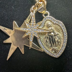 May include: Gold-toned pendant featuring a starburst design, a star with embedded crystals, and a heart-shaped charm with a religious figure. The pendant is suspended from a gold chain, creating a layered effect. The jewelry is set against a dark background.