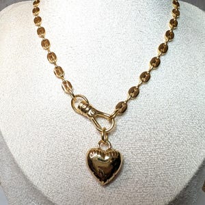 May include: A gold-colored necklace featuring a heart pendant. The necklace has a chain of small, round links and a lobster clasp. The heart pendant is a solid, rounded shape, and the necklace is displayed on a white surface.