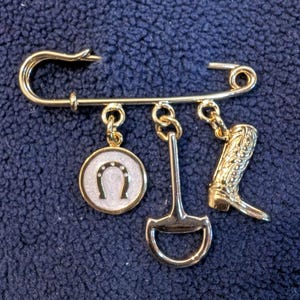 May include: Gold-toned safety pin with four charms: a horseshoe, a stirrup, a boot, and a round charm with a white enamel horseshoe design.