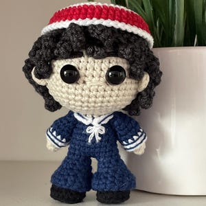 Op de afbeelding: Een handgemaakte gehaakte pop met donker krullend haar, een rood-witte pet en een marineblauw matrozenpakje met witte randen en een kleine witte strik bij de kraag. De pop heeft grote zwarte plastic ogen en een lichtbeige garen huid.