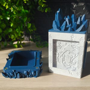 Op de afbeelding: Tweedelige set decoratieve containers. Eén is een rechthoekige, diepblauwe doos met een grillige, kristalachtige rand. De andere is een witte en grijze rechthoekige doos met een blauwe kristalachtige bovenkant en een gebeeldhouwd berenontwerp.