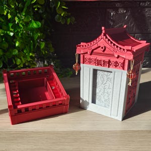 Op de afbeelding: Een rode, miniatuur pagode-achtige doos met een bijpassend rood deksel. De doos heeft een wit voorpaneel met een reliëf ontwerp. Decoratieve rode lantaarns hangen aan het dak. De doos staat op een houten oppervlak.