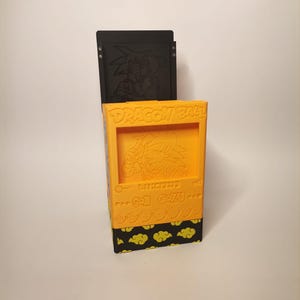 Puede incluir: Una caja de almacenamiento de color amarillo y negro con tem&aacute;tica de Dragon Ball Z con una imagen en relieve de Goku y el texto "Dragon Ball Z" y "Son Goku".