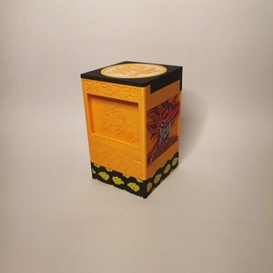 Puede incluir: Una caja de cartas Dragon Ball Z impresa en 3D de color amarillo y negro con un s&iacute;mbolo de bola de drag&oacute;n en la parte superior y una imagen de Goku en la parte delantera.