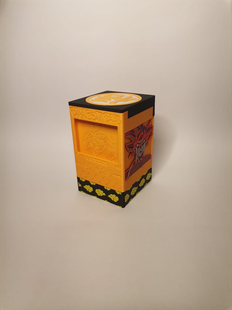Puede incluir: Una caja de cartas Dragon Ball Z impresa en 3D de color amarillo y negro con un s&iacute;mbolo de bola de drag&oacute;n en la parte superior y una imagen de Goku en la parte delantera.