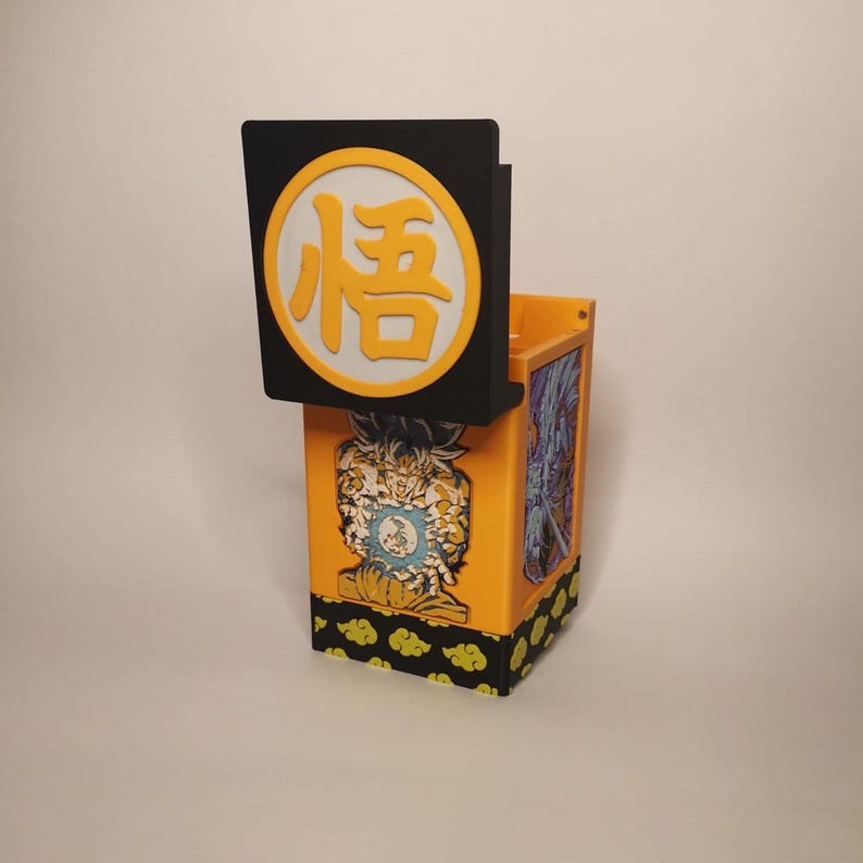 Puede incluir: Una caja impresa en 3D de color amarillo y negro con un personaje del anime Dragon Ball Z en la parte delantera. La caja tiene una tapa con fondo negro y un c&iacute;rculo amarillo con el kanji para "suerte" en blanco. La caja est&aacute; decorada con un patr&oacute;n de nubes amarillas sobre un fondo negro.