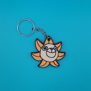 May include: A yellow and white cartoon sun keychain with a smiling face. The keychain has a silver metal ring.