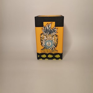 Puede incluir: Una caja amarilla y negra con un gr&aacute;fico de un personaje del anime Dragon Ball Z. El personaje est&aacute; en una pose de lucha con una esfera de energ&iacute;a azul en su mano. La caja tiene una base negra y una parte superior amarilla con un borde negro. La caja tiene un patr&oacute;n de nubes verdes en la parte inferior.
