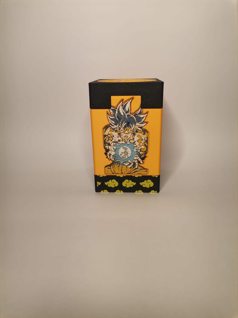 Puede incluir: Una caja amarilla y negra con un gr&aacute;fico de un personaje del anime Dragon Ball Z. El personaje est&aacute; en una pose de lucha con una esfera de energ&iacute;a azul en su mano. La caja tiene una base negra y una parte superior amarilla con un borde negro. La caja tiene un patr&oacute;n de nubes verdes en la parte inferior.