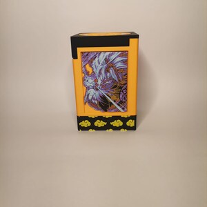 Puede incluir: Una caja amarilla y negra con una imagen impresa de un personaje de una serie de anime popular. La caja tiene un borde negro y un patr&oacute;n de nubes amarillas en la parte inferior.