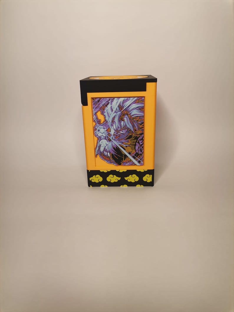 Puede incluir: Una caja amarilla y negra con una imagen impresa de un personaje de una serie de anime popular. La caja tiene un borde negro y un patr&oacute;n de nubes amarillas en la parte inferior.
