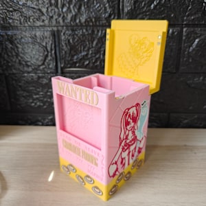 Op de afbeelding: Een roze en gele doos met een scharnierend deksel, met illustraties in anime-stijl en het woord "WANTED". De doos heeft een gele basis met cirkelvormige ontwerpen en een geel deksel met een omtrek van een personage. De doos is versierd met anime-personages.