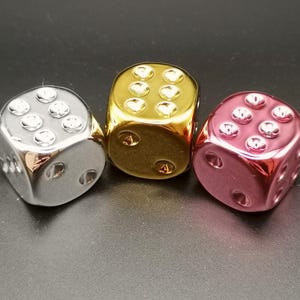 Puede incluir: Tres dados met&aacute;licos en plata, oro y rosa. Los dados son todos del mismo tama&ntilde;o y forma, y est&aacute;n todos sobre una superficie negra.