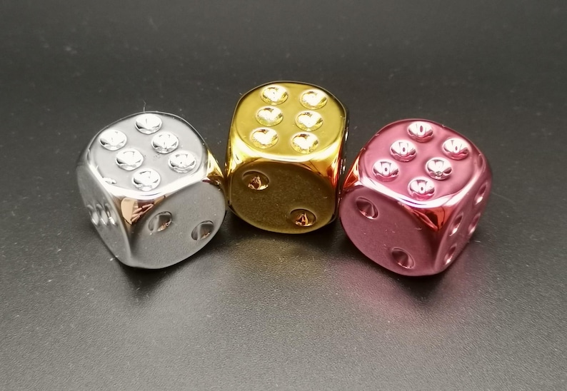 Puede incluir: Tres dados met&aacute;licos en plata, oro y rosa. Los dados son todos del mismo tama&ntilde;o y forma, y est&aacute;n todos sobre una superficie negra.