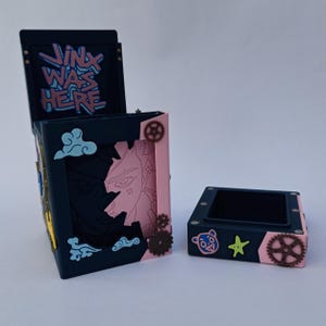 Op de afbeelding: Een donkerblauwe doos met de tekst "Jinx Was Here" in lichtblauw. De doos heeft een uitgesneden ontwerp met een roze interieur en tandwielaccenten. Een kleinere bijpassende doos is ook aanwezig, met een beer en een ster.