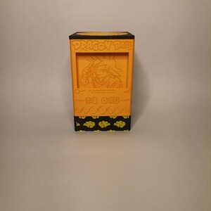 Puede incluir: Caja naranja con tem&aacute;tica de Dragon Ball Z con tapa negra y una imagen de Son Goku en la parte delantera. La caja tiene un patr&oacute;n de nubes amarillas sobre un fondo negro.
