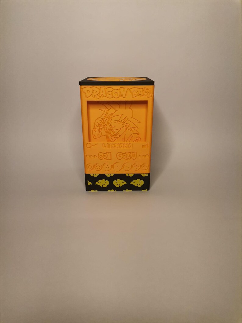 Puede incluir: Caja naranja con tem&aacute;tica de Dragon Ball Z con tapa negra y una imagen de Son Goku en la parte delantera. La caja tiene un patr&oacute;n de nubes amarillas sobre un fondo negro.