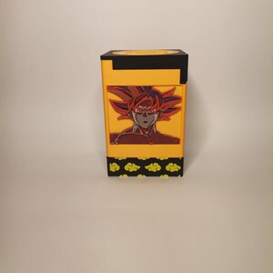 Puede incluir: Una caja amarilla y negra con una imagen roja y naranja de un personaje con el pelo de punta y ojos rojos. La caja tiene un patr&oacute;n de nubes amarillas sobre un fondo negro.
