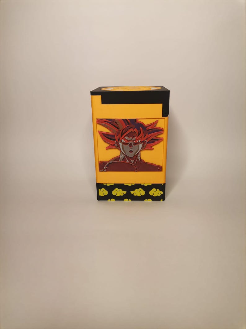 Puede incluir: Una caja amarilla y negra con una imagen roja y naranja de un personaje con el pelo de punta y ojos rojos. La caja tiene un patr&oacute;n de nubes amarillas sobre un fondo negro.