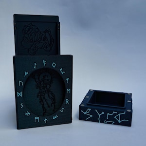 Op de afbeelding: Een donkerblauwe doosset met een scharnierend deksel en een aparte kleine doos. De grotere doos heeft een cirkelvormig reliëf van een gevleugelde figuur en runen. Het deksel en de kleine doos hebben geëtste ontwerpen.