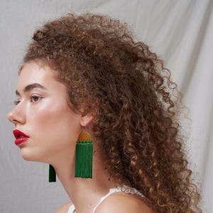 以下が含まれることがあります： 白いタンクトップを着て、金色のアクセントがついた緑色のタッセルイヤリングをしている、巻き毛の茶髪の女性です。