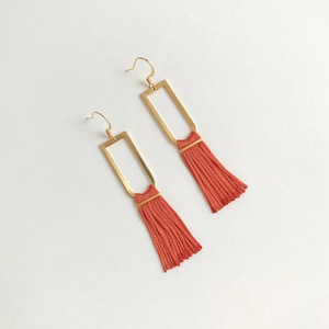 以下が含まれることがあります： 長方形のフレームと長いオレンジ色のタッセルが付いた、ゴールドのイヤリングのペアです。