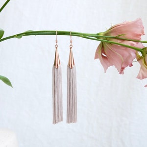以下が含まれることがあります： 金色の先端を持つ、長い銀色のタッセルイヤリングのペア。イヤリングは、ピンクの花が咲いた茎からぶら下がっています。