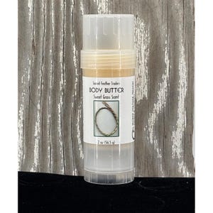 May include: A clear plastic tube of body butter with a white label. The label reads "Sacred Feather Traders BODY BUTTER Sweet Grass Scent" and includes a graphic. The tube contains 2 oz (56.5 g) of product.