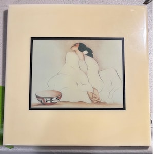 May include: A square ceramic tile featuring a centered illustration of a person with dark hair tied back, wearing a flowing white garment, sitting beside a small bowl decorated with geometric black and red patterns on a cream-colored background.