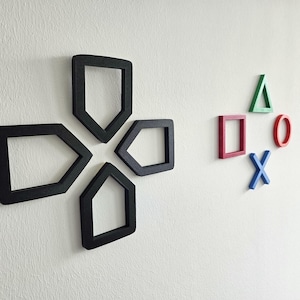 Könnte beinhalten: Schwarz-weißes PlayStation-Controller-Symbol aus Holz, an einer weißen Wand montiert. Daneben befinden sich die Symbole für die PlayStation-Tasten: Dreieck, Quadrat, Kreis und X in Grün, Rot, Rot und Blau.