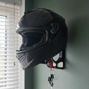 May include: A glossy black motorcycle helmet mounted on a black wall bracket. The helmet is hanging on a wall with a red lanyard and keys attached. The keys have a Mercedes-Benz logo.