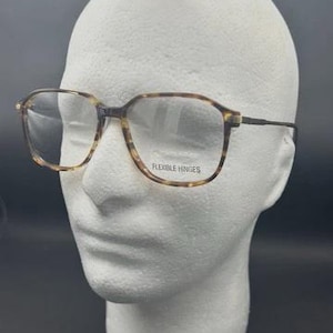 May include: Tortoiseshell pattern eyeglasses with clear lenses on a white mannequin head. The glasses have a rectangular shape with rounded edges. The text "FLEXIBLE HINGES" is visible on the frame. The frame is brown and tan.