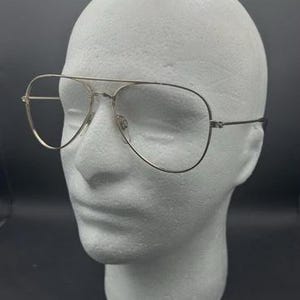 Könnte beinhalten: Pilotenbrille mit einem dünnen, goldfarbenen Metallrahmen. Die Brille hat einen Doppelsteg und tropfenförmige Gläser. Die Brille wird auf einem weißen Schaumstoffkopf vor dunklem Hintergrund präsentiert.