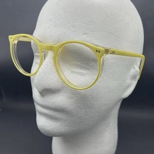 May include: Pale yellow round eyeglasses with clear lenses are displayed on a white mannequin head. The frames have a subtle, translucent quality, and the overall design is simple and modern. The glasses are a soft, pastel yellow.